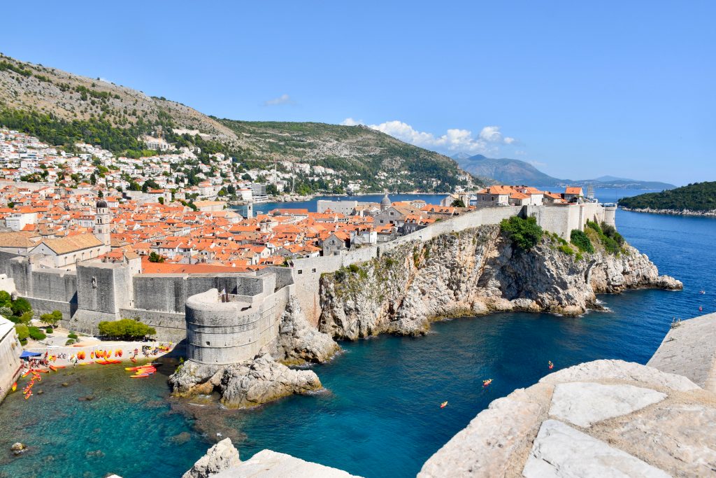 A walled city sits on rocky cliffs from the left hand side to the middle of the frame. The centre of the Old Town is crammed with tiled orange roofed buildings and some church towers. Hills behind. Water around the right hand side and a small pebble beach filled with orange kayaks to the bottom left