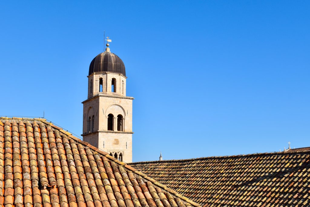 View across terracotta roof tiles to a square tower with domed roof. Clear blue sky beyond.