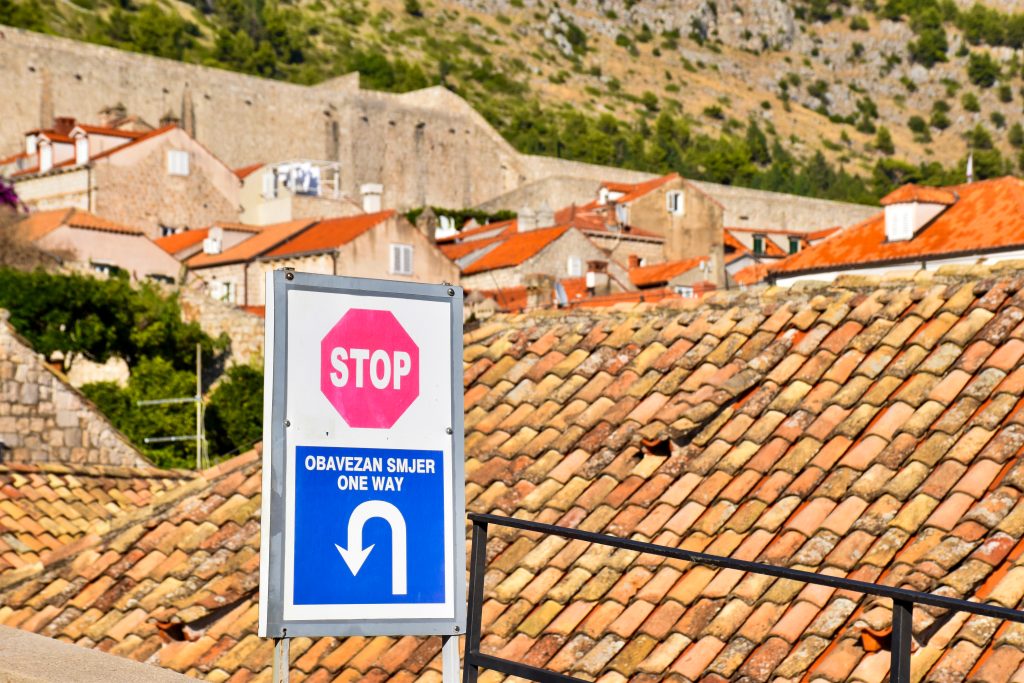A white sign post sits to the middle left of the shot. It has a red stop sign on it and a blue notice saying 'One Way'. In the background are terracotta tiles on top stone buildings. Rocky hill behind.