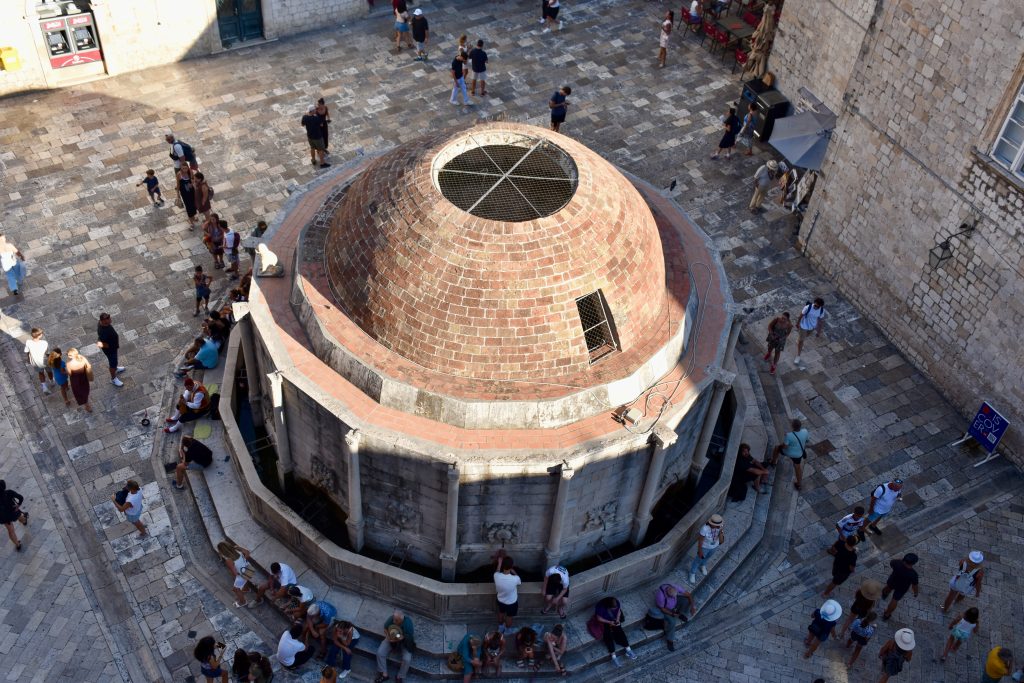 Looking down on an impressive circular building with domed roof. The structure is large and las steps all the way around, with people sitting on the steps. Lots of people are milling around. The structure appears to sit in a square