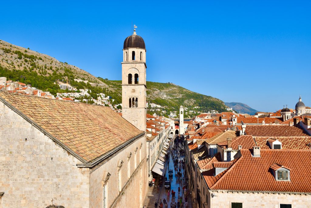Looking down a narrow street with impressive buildings on either side. To the left is a stone church or monastery building with a tall domed tower. To the right are buildings with red roofs. Pedestrians on the streets. Hills beyond