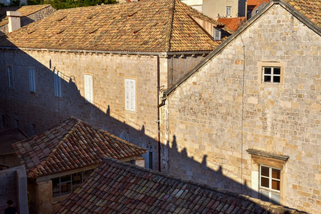 View across to tall stone walls beneath terracotta roof tiles. A diagonal shadow is cast on the buildings opposite showing people walking along a wall in silhouette