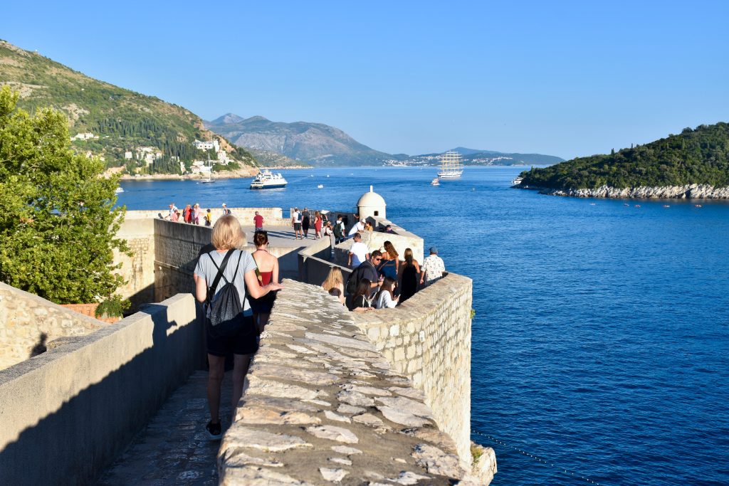 Following a line of people walking along Old City stop walls towards a viewpoint platform at the end. To the right is a body of water with a couple of ships on it - one a sailing ship with masts. To the right is a stony island covered in green trees. Hills to the left