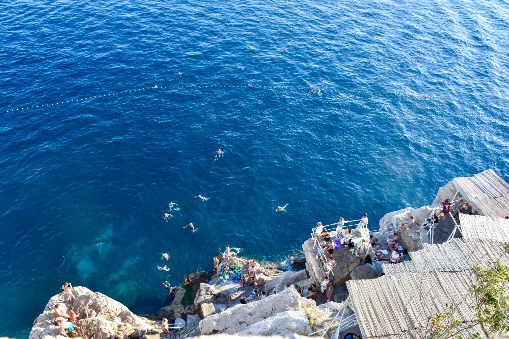 Looking down on clear blue water from the top of a tall cliff or wall. It appears that there is a small bar attached to the side of the cliff with people standing or sitting, watching swimmers in the water below