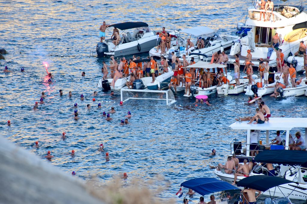 View looking down on a water scene. It looks like a game of water polo in the sea with two teams of swimmings, some in red caps and some in blue. Across the top and to the right of the swimmers are rows of small moored boats filled with observers standing or sitting and watching the game, many of them in orange t-shirts