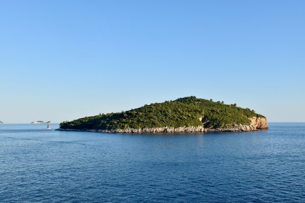 View of a small rocky island covered in trees. Surrounded by calm sea and blue sky above. One small boat to the left of the island