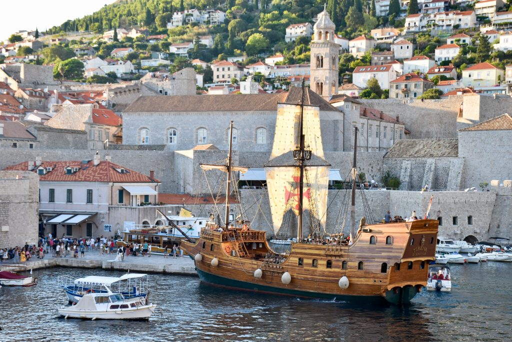 View of a brown galleon ship with tall cream sails moored in port with old stone fortress buildings behind. A hill covered in white houses with terracotta roofs behind