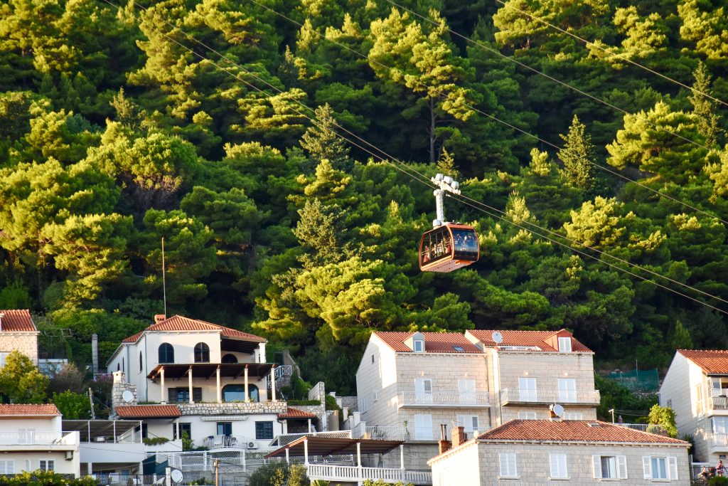 View of a red cable car passing across the middle of the frame on diagonal wires. Trees behind. Terracotta roofs on stone buildings below
