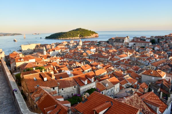 Picture postcard view over an old walled city filled with terracotta roofs and church towers in the glow of sunset. Beyond is a calm sea and then a small rocky tree covered island. Pale blue sky with pink and yellow shading on the horizon