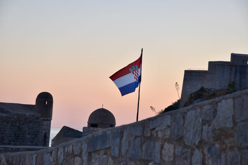 View of a flag flying in the centre of the frame above an old stone wall. Behind is an orange glow of dying sunset.