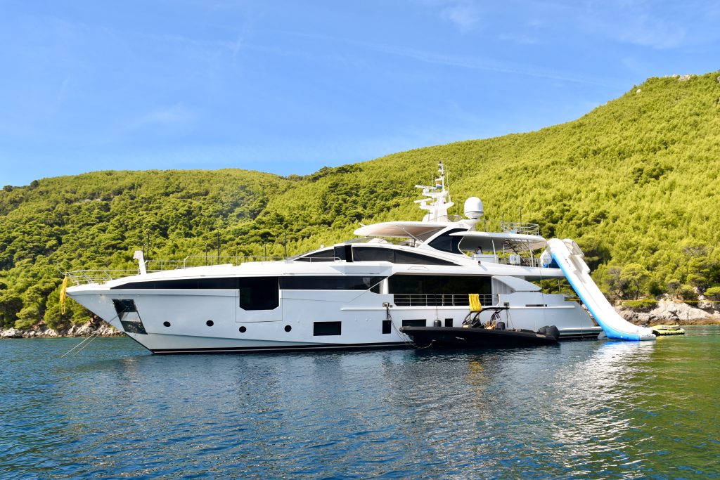 A large white super yacht is moored across the centre of the frame. The boat has a small black speed boat moored alongside and has a slide off the back of the boat. Still water in front. Trees behind. Pale blue sky above