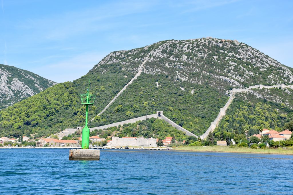 View across water to a rocky hill covered in trees. It also features an old city wall in a W shape. Green life buoy in the water to the left