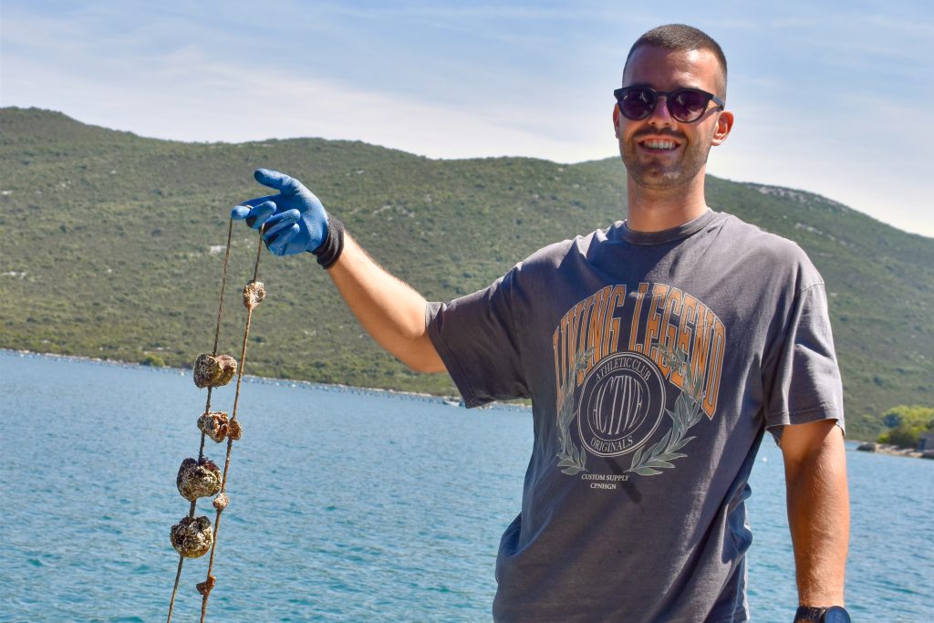 Man in navy blue t-shirt holds up a string of oysters up to the left showing them to the camera. He has a blue plastic glove on his hand, is wearing sunglasses and has an orange Legend logo on the front. Green hill behind.