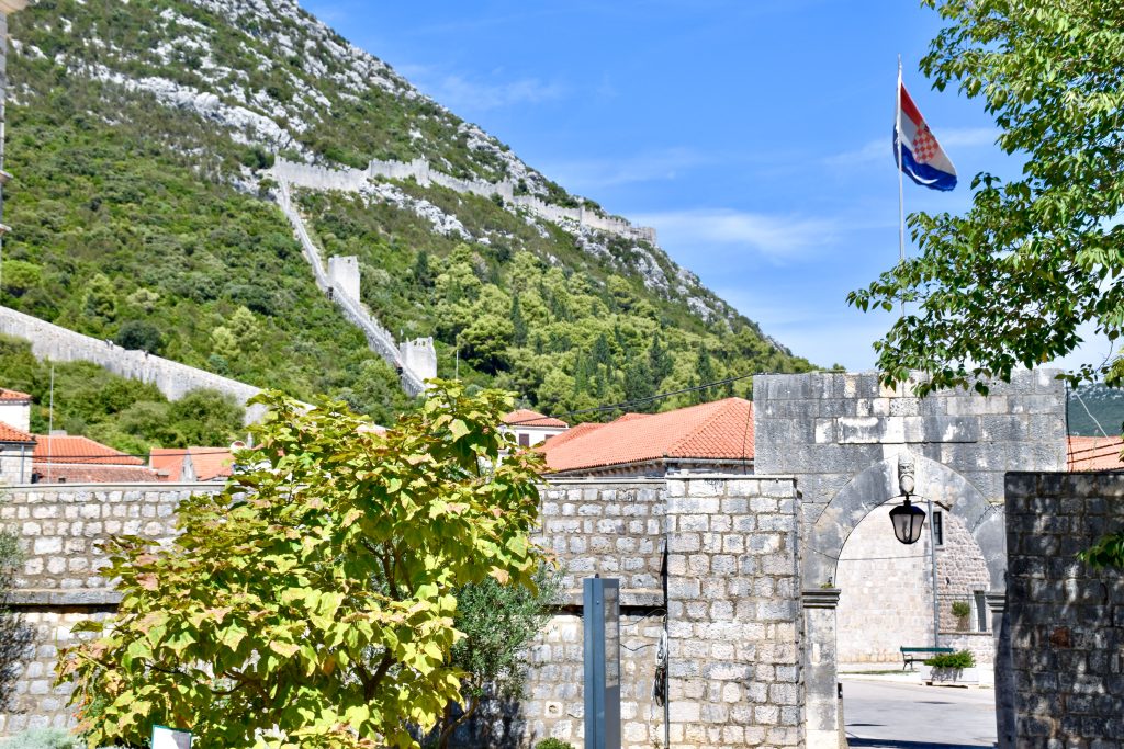 An old wall stretches across the bottom of the frame with an arched doorway to the right. Behind to the left are hills with zig zag city walls with turrets. Pale blue sky