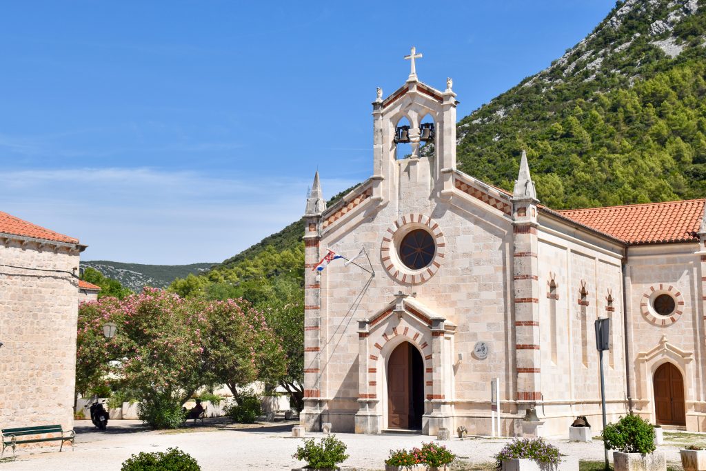 A white stone building with red stripes sits to the right hand side. It has an exposed bells at the top. Green hill behind. Terracotta roof.