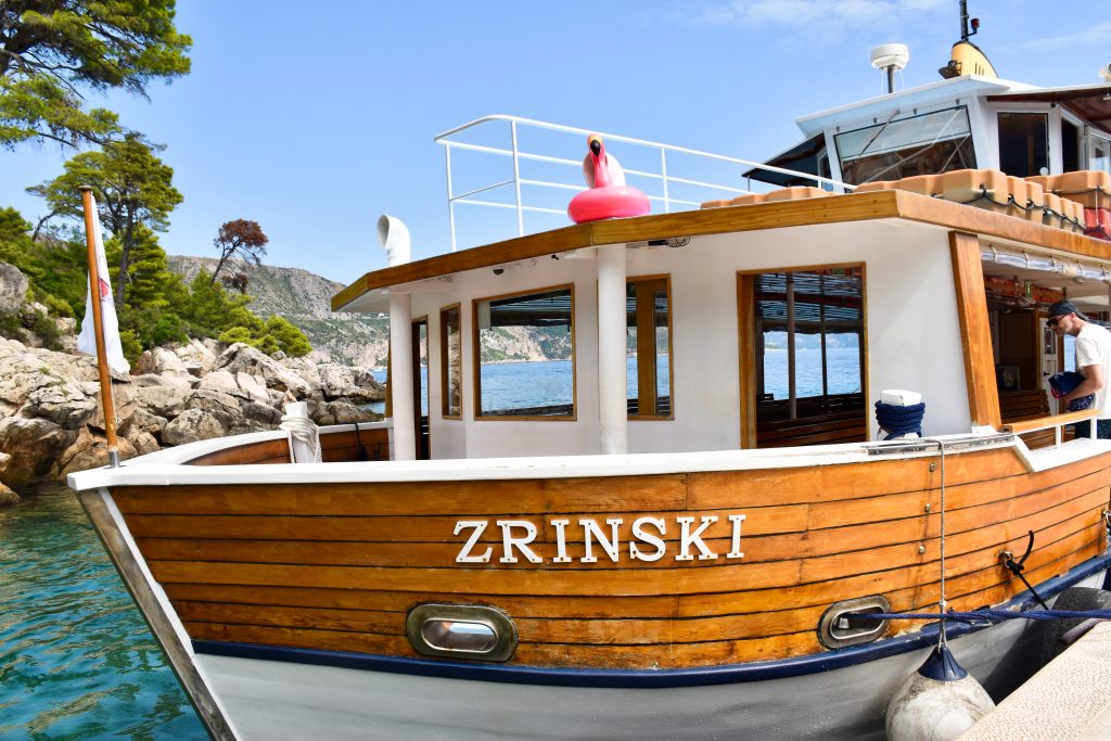 A wooden and white ferry boat fills the frame with rocky island behind. The boat says Zrinkski on the side and has a pink blow up flamingo on the roof