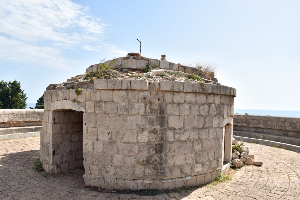 A circular stone structure with entrance way to the left and a window to the right. Bricks and rubble on top. It stands on a circular walled patio. Pale blue sky beyond