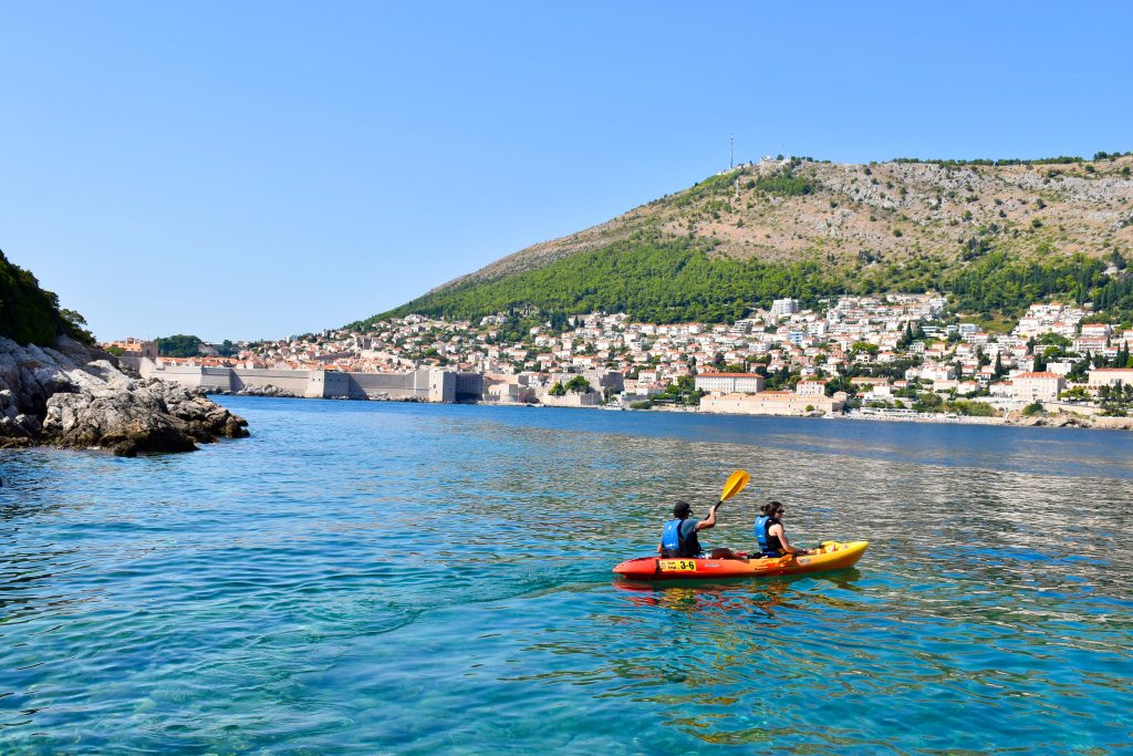 View over a calm body of turquoise water to a walled city. Small stone buildings with terracotta roofs on the hill above the old walls. Pale blue sky above. A red and yellow kayak in the foreground.
