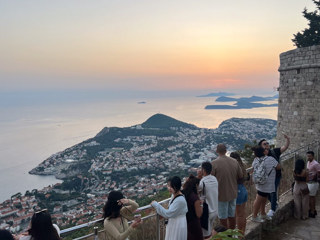 Sunset view from a balcony overlooking a coastal town below - mix of trees and terracotta roof tiles. Row of tourists on the balcony admiring the view. Small islands to the right hand side