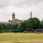 Looking across a grassy field to a row of trees with a green bridge in front. Behind the trees you can see the tops of a series of brick buildings and chimneys which look like those of a mill. Grey sky above
