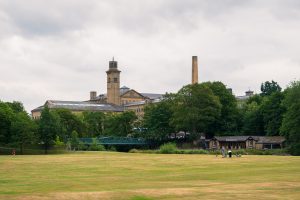 Looking across a grassy field to a row of trees with a green bridge in front. Behind the trees you can see the tops of a series of brick buildings and chimneys which look like those of a mill. Grey sky above