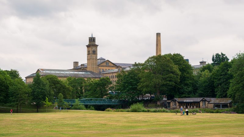 Looking across a grassy field to a row of trees with a green bridge in front. Behind the trees you can see the tops of a series of brick buildings and chimneys which look like those of a mill. Grey sky above
