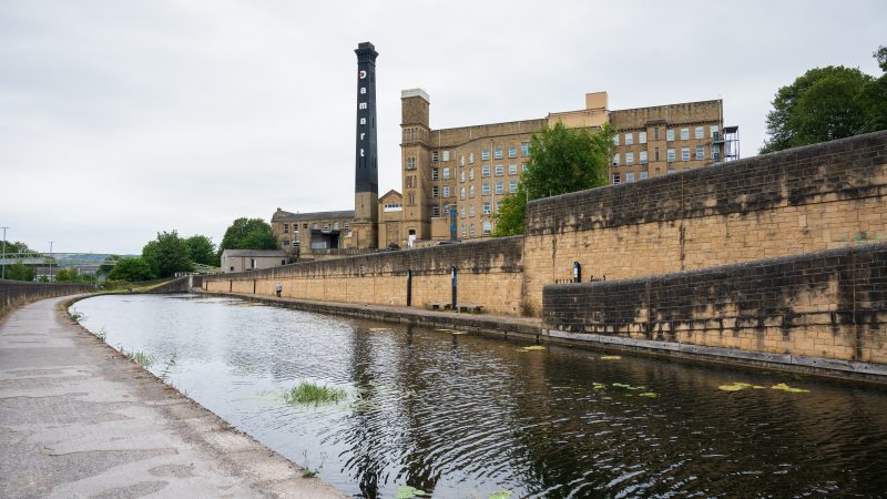 A canal leads from the bottom right to the middle left. Tow path to the left of the canal. Stone wall on the right hand side with a large old mill building to the right hand side. A stone chimney painted black at the top stands to the left of the mill. It says Damart on it