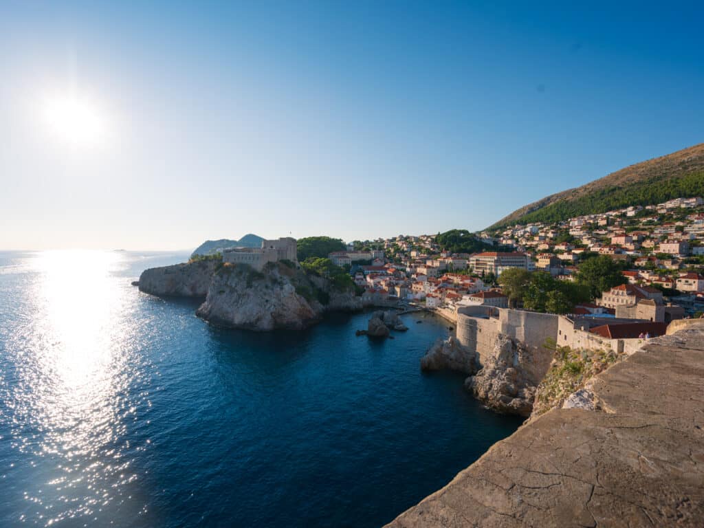 Coastal panorama of Dubrovnik showing a stone fortress perched on a rocky cliff above sparkling blue sea, with the old town’s red-tiled roofs climbing a green hillside under a clear sky and low sun.