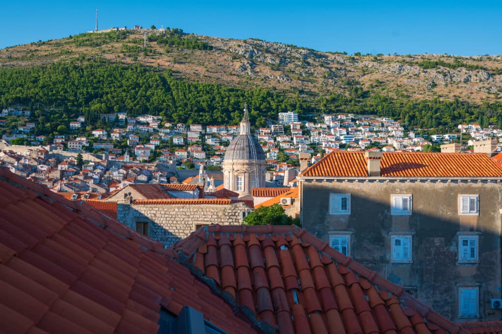 Rooftop view across Dubrovnik’s red-tiled roofs toward a church dome and the steep green mountain rising behind the city under a bright blue sky.