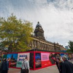 View towards a large Town Hall looking Victorian building with red billboards in front of it which looks like it contains construction site. People are walking on the street in front. The building is long and columned and has a tall clock tower with domed top.