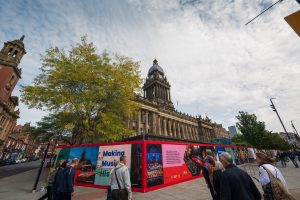 View towards a large Town Hall looking Victorian building with red billboards in front of it which looks like it contains construction site. People are walking on the street in front. The building is long and columned and has a tall clock tower with domed top.