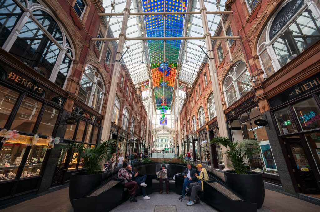 Looking into an up market shopping centre with shops on both sides. Glass roof with coloured glass at the top of the roof. At the bottom is a horseshoe shaped seat with 5 shoppers relaxing together
