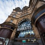 Looking up at a Victorian looking stone building with central glass doorway and sign saying Leeds City Markets in white. Brown curved designs either side of the doorway