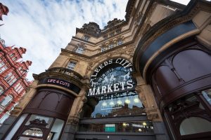 Looking up at a Victorian looking stone building with central glass doorway and sign saying Leeds City Markets in white. Brown curved designs either side of the doorway