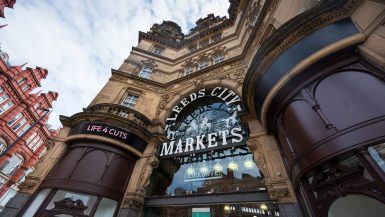 Looking up at a Victorian looking stone building with central glass doorway and sign saying Leeds City Markets in white. Brown curved designs either side of the doorway