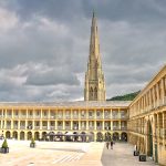 Looking into a corner of a piazza with columned sides along the right and straight in front. The building has three storeys with arches along the bottom and then windows on the two two but with columns in front indicating covered walkways around the building. A spire appears behind. Grey sky above