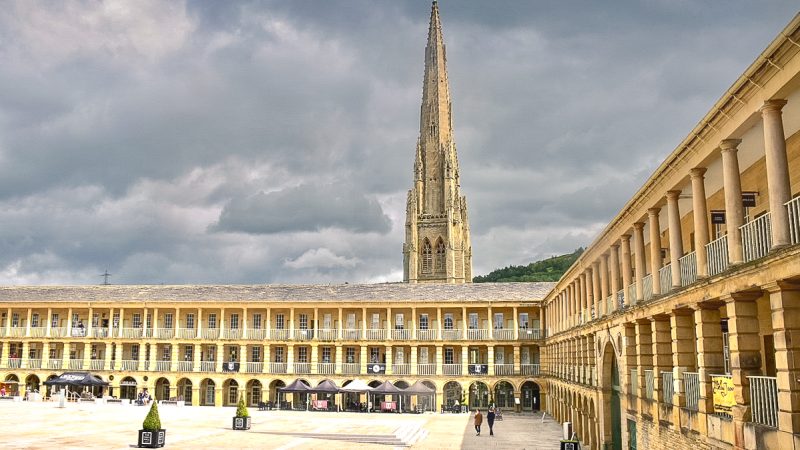 Looking into a corner of a piazza with columned sides along the right and straight in front. The building has three storeys with arches along the bottom and then windows on the two two but with columns in front indicating covered walkways around the building. A spire appears behind. Grey sky above