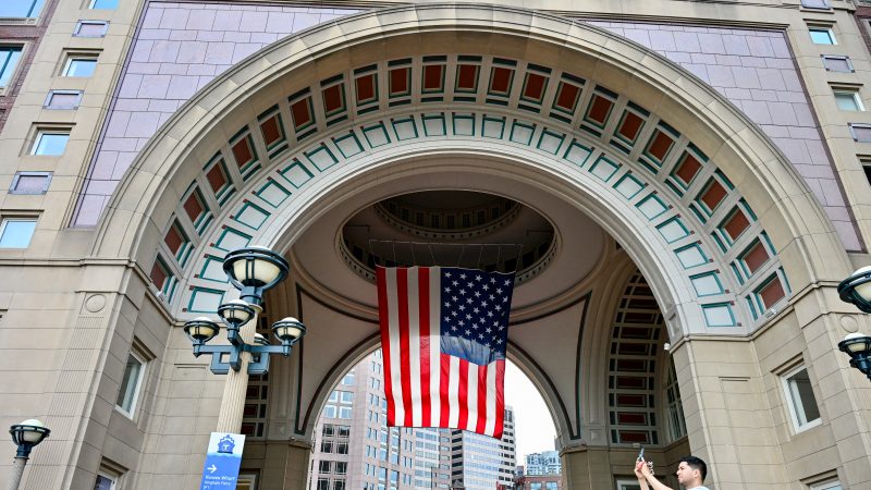 A large archway is built into a modern building which is flying the Stars and Stripes from its ceiling. A man in the foreground takes a photo on his mobile phone