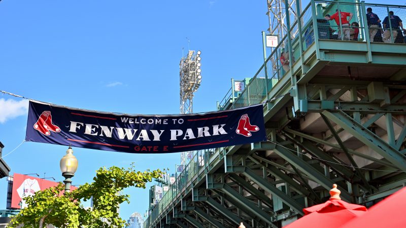 Looking up at the back of a green stadium. Blue sky above and a navy blue banner across the walkway which says 'Welcome to Fenway Park, GatecE'