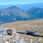 View from a rocky hill down to a rocky hill top with a pink railway carriage making its way from left to right across the frame. Beyond the hill top are the tops of mountains. Beyond that are pale hills behind.