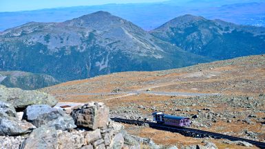 View from a rocky hill down to a rocky hill top with a pink railway carriage making its way from left to right across the frame. Beyond the hill top are the tops of mountains. Beyond that are pale hills behind.