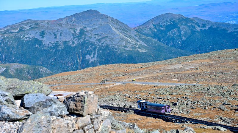 View from a rocky hill down to a rocky hill top with a pink railway carriage making its way from left to right across the frame. Beyond the hill top are the tops of mountains. Beyond that are pale hills behind.