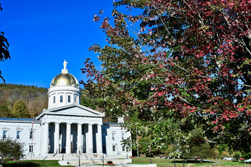 A white stone building with gold dome roof stands to the left of the frame. It looks like a mini White House. Hils and blue sky behind. Trees with some red leaves to the right