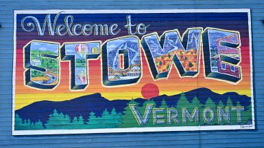 A blue painted wooden clapboard wall with a painted mural saying 'Welcome to Stowe, Vermont'. Each letter is filled with an outdoor scene. Above a line of mountains, fir trees and sunset scene