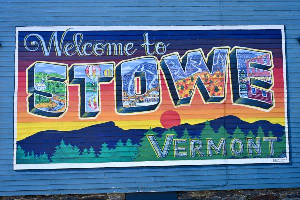 A blue painted wooden clapboard wall with a painted mural saying 'Welcome to Stowe, Vermont'. Each letter is filled with an outdoor scene. Above a line of mountains, fir trees and sunset scene