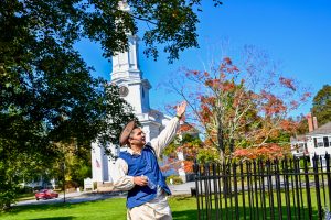 A man in period costume stands in the bottom middle of the frame pointing to something to his right. He is wearing white with a brown tricorn hat and blue waistcoat. He has a mouthpiece as if he's a tour guide. White church and trees behind.