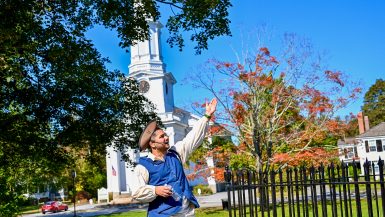 A man in period costume stands in the bottom middle of the frame pointing to something to his right. He is wearing white with a brown tricorn hat and blue waistcoat. He has a mouthpiece as if he's a tour guide. White church and trees behind.