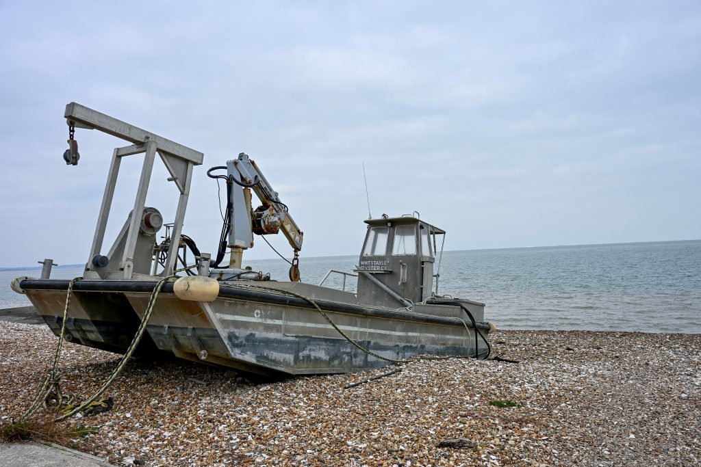 A grey flat bottomed oyster catcher boat sits on a pebble beach with grey sea and grey sky behind