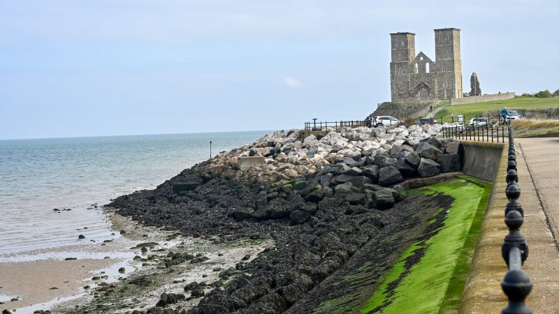 View along a concrete promenade to the right hand side towards a stone structure of two square towers joined by a wall with pointed roof. There is a black railing along the promenade and rocks and green moss to the left leading to a grey sea