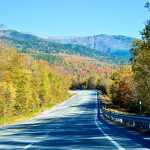 Looking down a tarmac road with white and yellow markings. It has a barrier to the right hand side and trees to the left and right. Mountains in the distance with multi-coloured leaves - greens, yellows and oranges as if Autumn